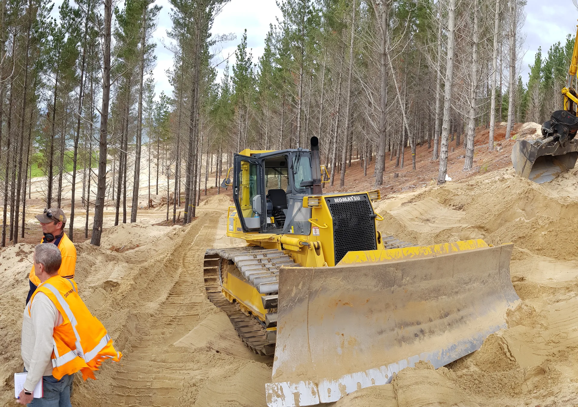Site manager overseeing earthworks — bulldozer and excavator working in a sandy cut with pine forest backdrop